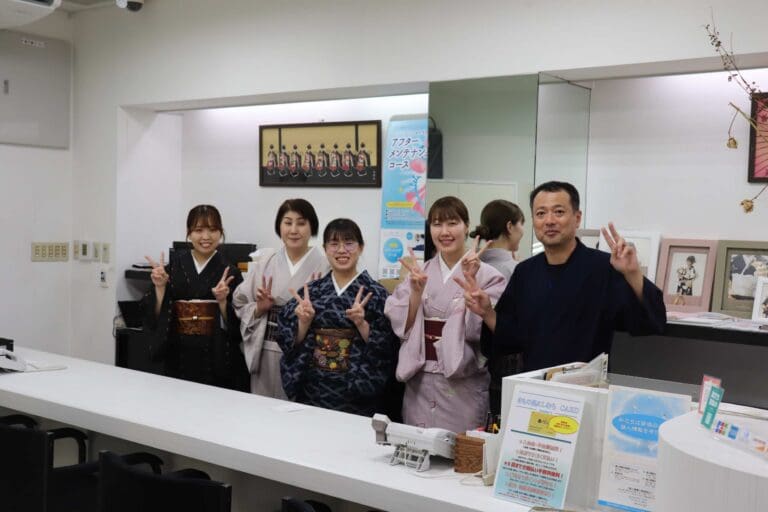 カウンターの後ろに5人が立っており、それぞれが手でピースサインをしています。彼らは日本の伝統的な衣装を着ています。背景にはポスターと案内パンフレットが展示されています。