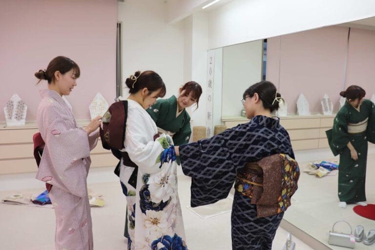 4 人の女性が部屋で伝統的な着物を試着している。3 人は笑顔で着物を整えており、緑色の服を着た 1 人の女性が鏡を見ている。部屋の壁は明るく、複数の着物とアクセサリーが見える。