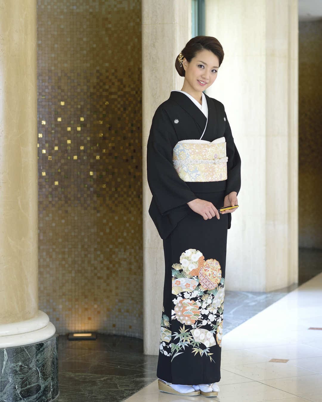 花柄の黒い着物と白と金の帯を締めた人物が室内に立っています。背景には大理石の床とベージュの柱があります。人物は小さな金色のクラッチバッグを持っています。