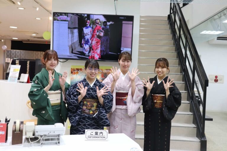 モダンな室内の階段の前に、伝統的な着物を着た 4 人が立っています。彼らは笑顔で手を挙げています。彼らの後ろには、着物を着た人の映像を映し出すテレビ スクリーンがあります。看板には「情報」と書かれています。
