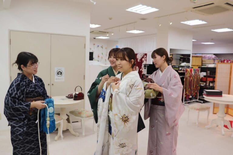 4 人の女性が明るい部屋にいます。そのうち 3 人が、4 人目の女性が花柄の白い着物を着るのを手伝っています。部屋には背景に着物があり、テーブルと椅子があります。