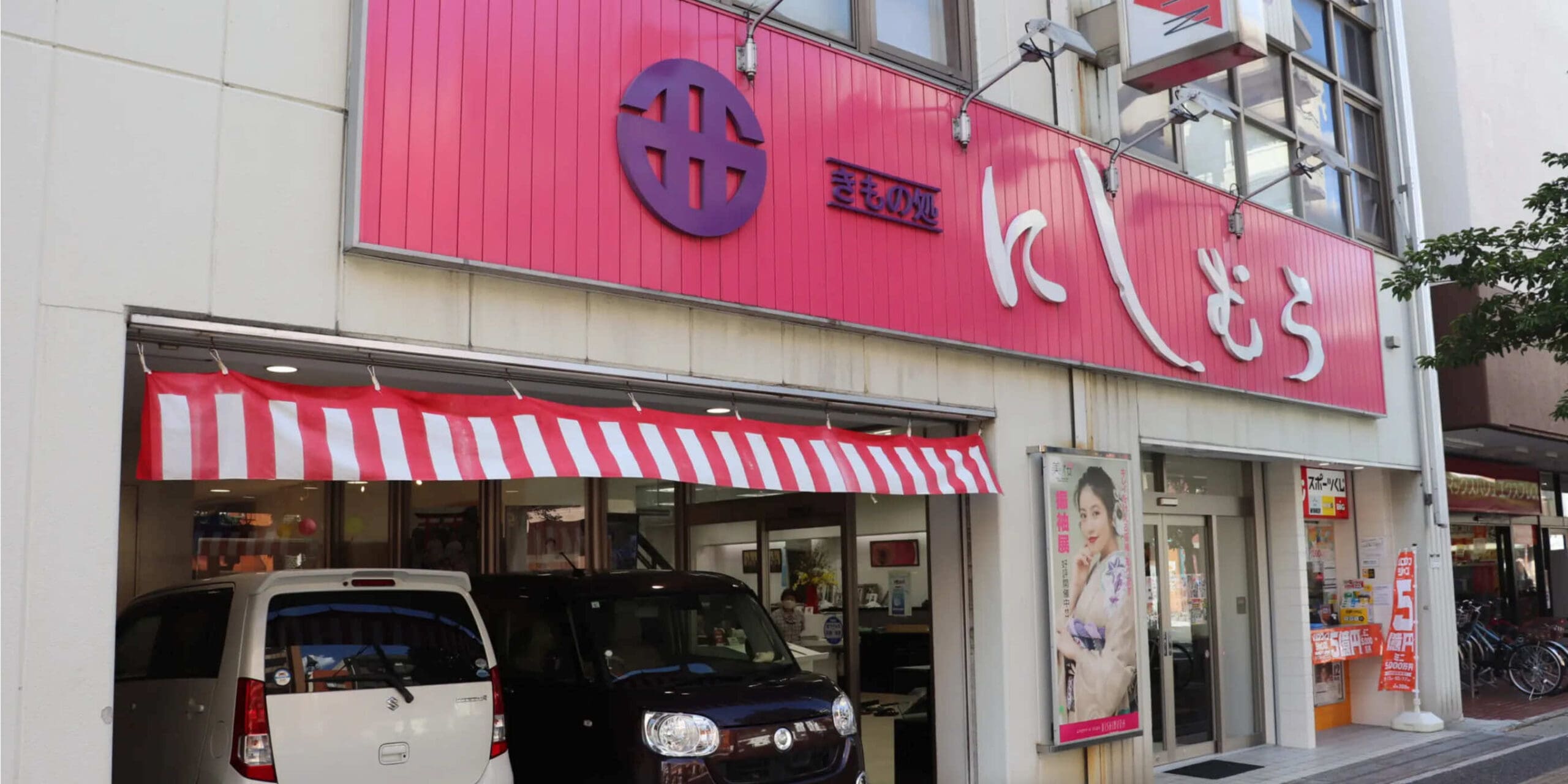 赤と白の縞模様のひさしと、白い日本語の文字が書かれた赤い看板のある店先。前には車が 2 台停まっています。窓にはポスターが貼られています。建物は複数階建てで、市街地にあります。