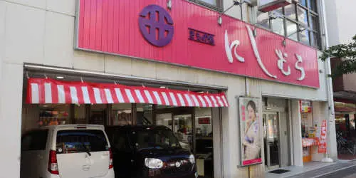 日本語の文字と円形のロゴが入ったピンクと赤の看板のある店先。店の前には車が 2 台停まっており、赤と白の縞模様の日よけがあります。入り口近くには人物が描かれたポスターが見えます。