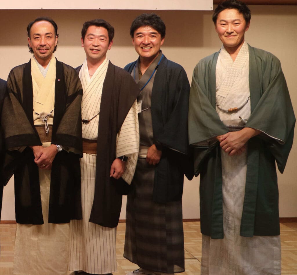 屋内に並んで立つ4人の男性が、着物や羽織などの伝統的な和服を着てカメラに微笑みかけている。無地の背景と木の床が落ち着いた雰囲気で、季節のイベントやイベント情報の告知にぴったりです。.