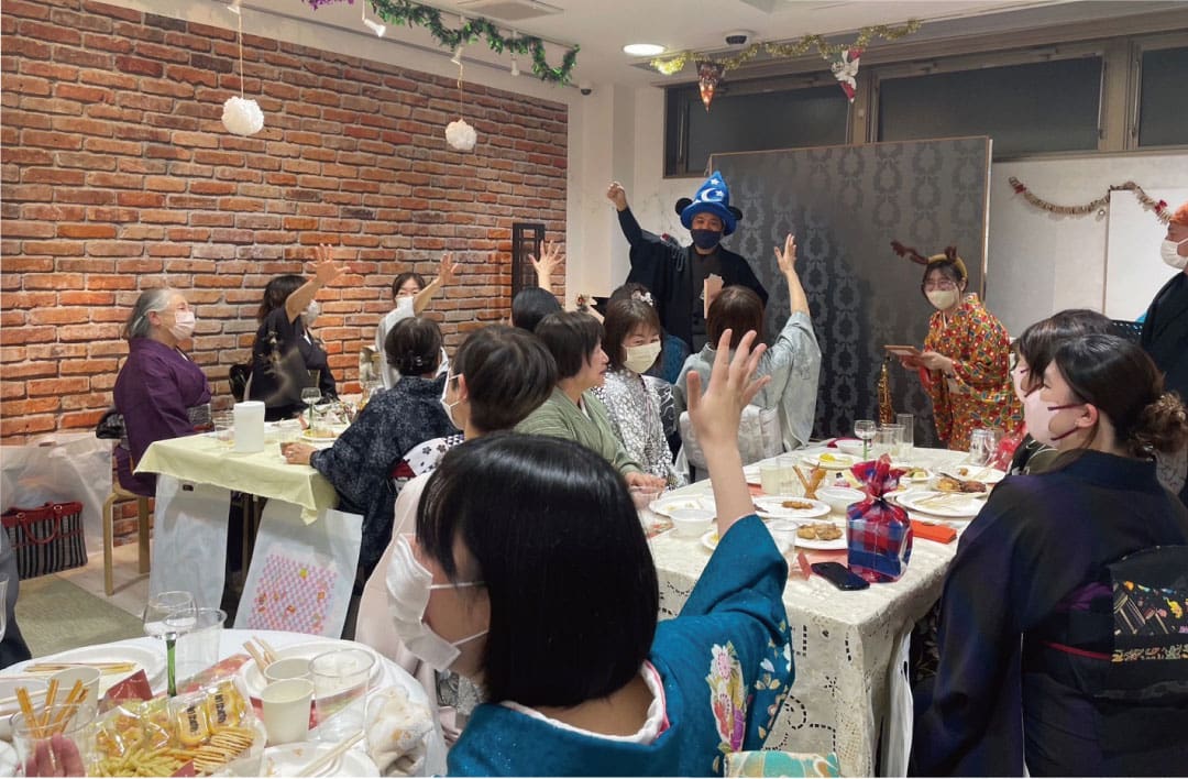 賑やかな着物イベントでは、仮面や着物姿の人々が、料理が並べられた飾り台の周りに集まる。魔法使いの帽子をかぶった一人の客が先頭で手を挙げ、他の客も加わり、祝祭的で優雅な雰囲気に包まれる。.